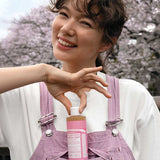 Smiling woman in pink overalls holding the cap of a pink-labelled Dr. Bronner's Cherry Blossom liquid soap bottle tucked into her front pocket.