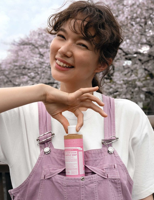 Smiling woman in pink overalls holding the cap of a pink-labelled Dr. Bronner's Cherry Blossom liquid soap bottle tucked into her front pocket.