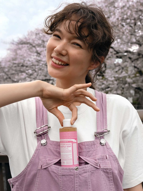Smiling woman in pink overalls holding the cap of a pink-labelled Dr. Bronner's Cherry Blossom liquid soap bottle tucked into her front pocket.