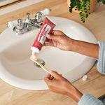 Hands squeezing red Dr. Bronner's Cinnamon Toothpaste onto a wooden toothbrush above a white bathroom basin.