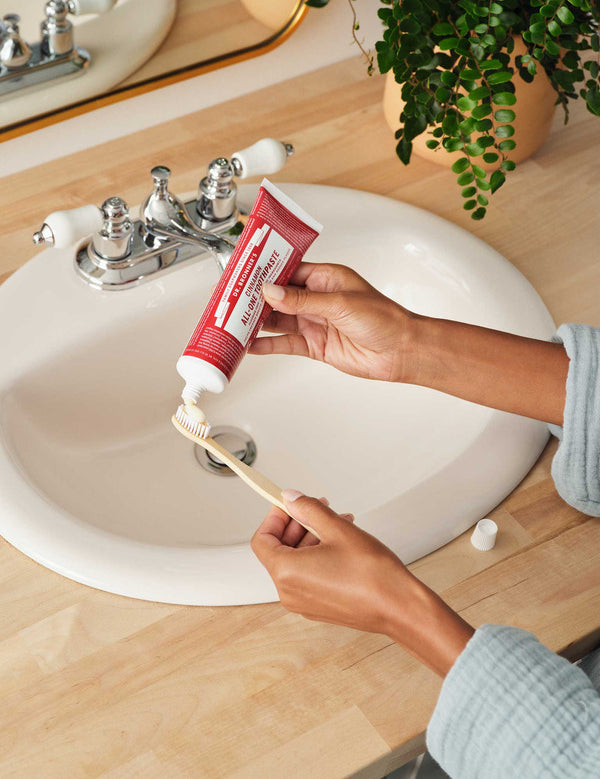 Hands squeezing red Dr. Bronner's Cinnamon Toothpaste onto a wooden toothbrush above a white bathroom basin.