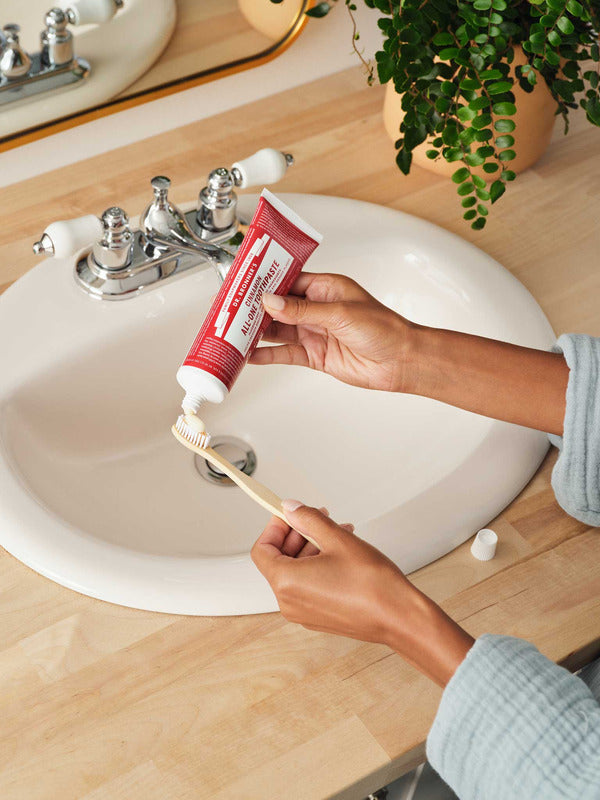 Hands squeezing red Dr. Bronner's Cinnamon Toothpaste onto a wooden toothbrush above a white bathroom basin.