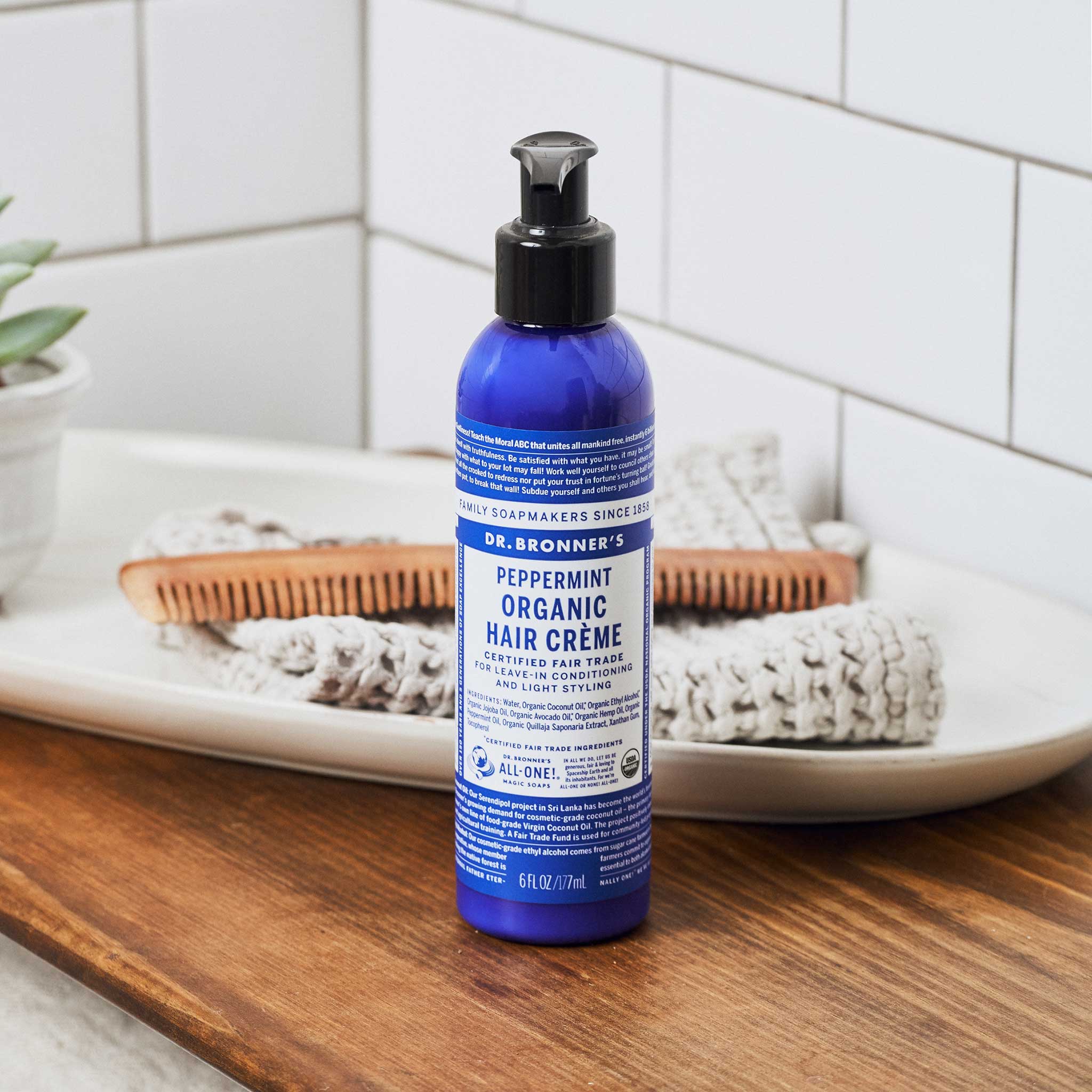Blue Dr. Bronner's Peppermint Hair Crème pump bottle on a wooden bathroom counter beside a white tray holding a folded towel and wooden comb.