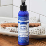 Dr. Bronner's Peppermint Leave In Conditioner bottle displayed on a bathroom counter with hair brushes.