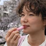 Model applying Dr. Bronner's Cherry Blossom Organic Lip Balm outdoors beneath flowering trees.