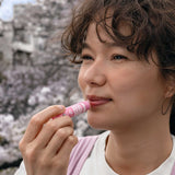 Model applying Dr. Bronner's Cherry Blossom Organic Lip Balm outdoors beneath flowering trees.