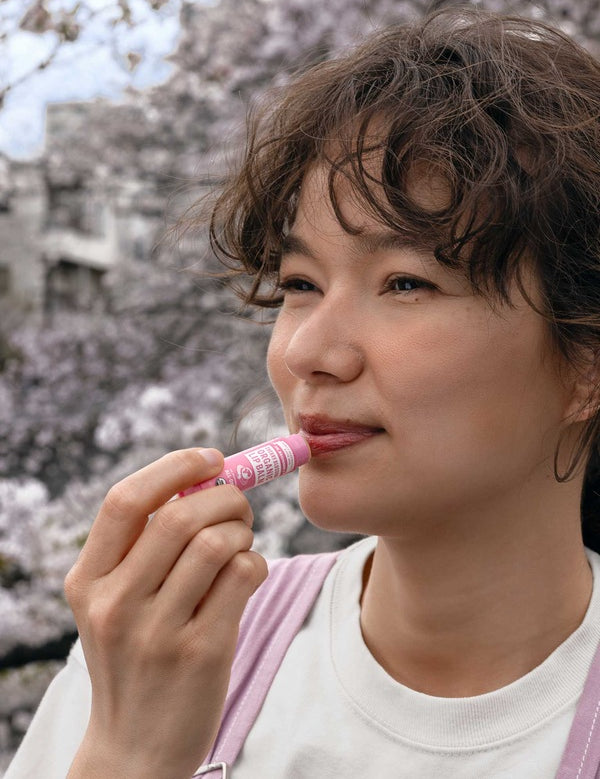Model applying Dr. Bronner's Cherry Blossom Organic Lip Balm outdoors beneath flowering trees.