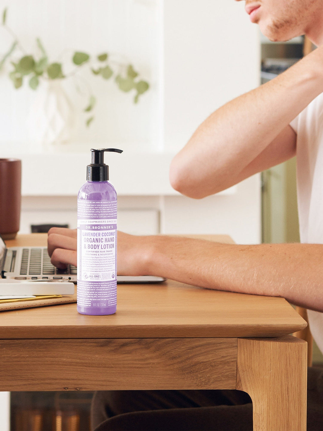 Person touching the back of their neck while seated at a wooden desk with a laptop, beside a purple Dr. Bronner's Lavender Coconut Organic Hand & Body Lotion 237ml pump bottle.