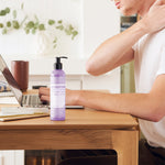 Person touching the back of their neck while seated at a wooden desk with a laptop, beside a purple Dr. Bronner's Lavender Coconut Organic Hand & Body Lotion 237ml pump bottle.