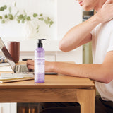 Person touching the back of their neck while seated at a wooden desk with a laptop, beside a purple Dr. Bronner's Lavender Coconut Organic Hand & Body Lotion 237ml pump bottle.