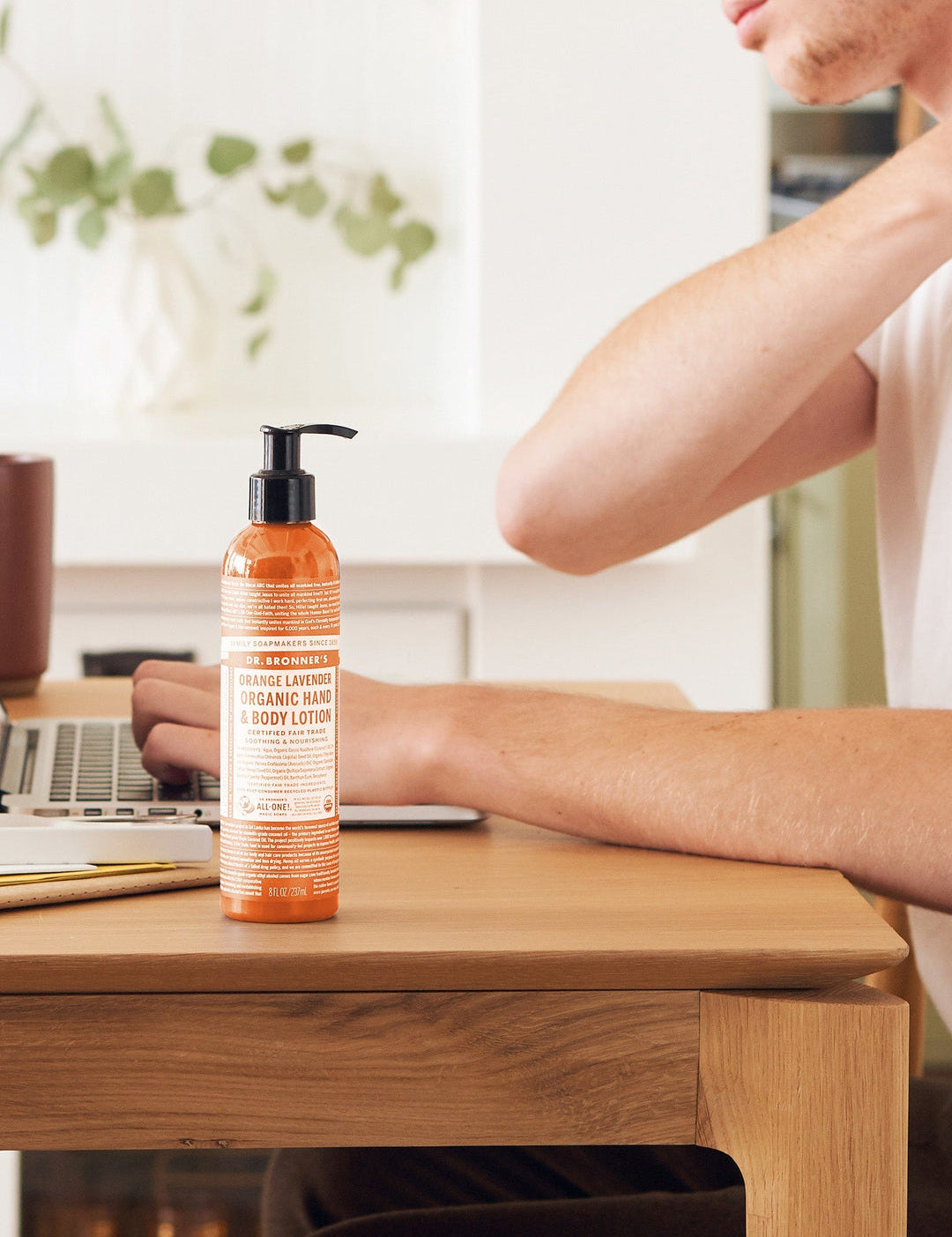 Person touching the back of their neck while seated at a wooden desk with a laptop, beside a orange Dr. Bronner's Orange Lavender Organic Hand & Body Lotion 237ml pump bottle.