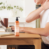 Person touching the back of their neck while seated at a wooden desk with a laptop, beside a orange Dr. Bronner's Orange Lavender Organic Hand & Body Lotion 237ml pump bottle.
