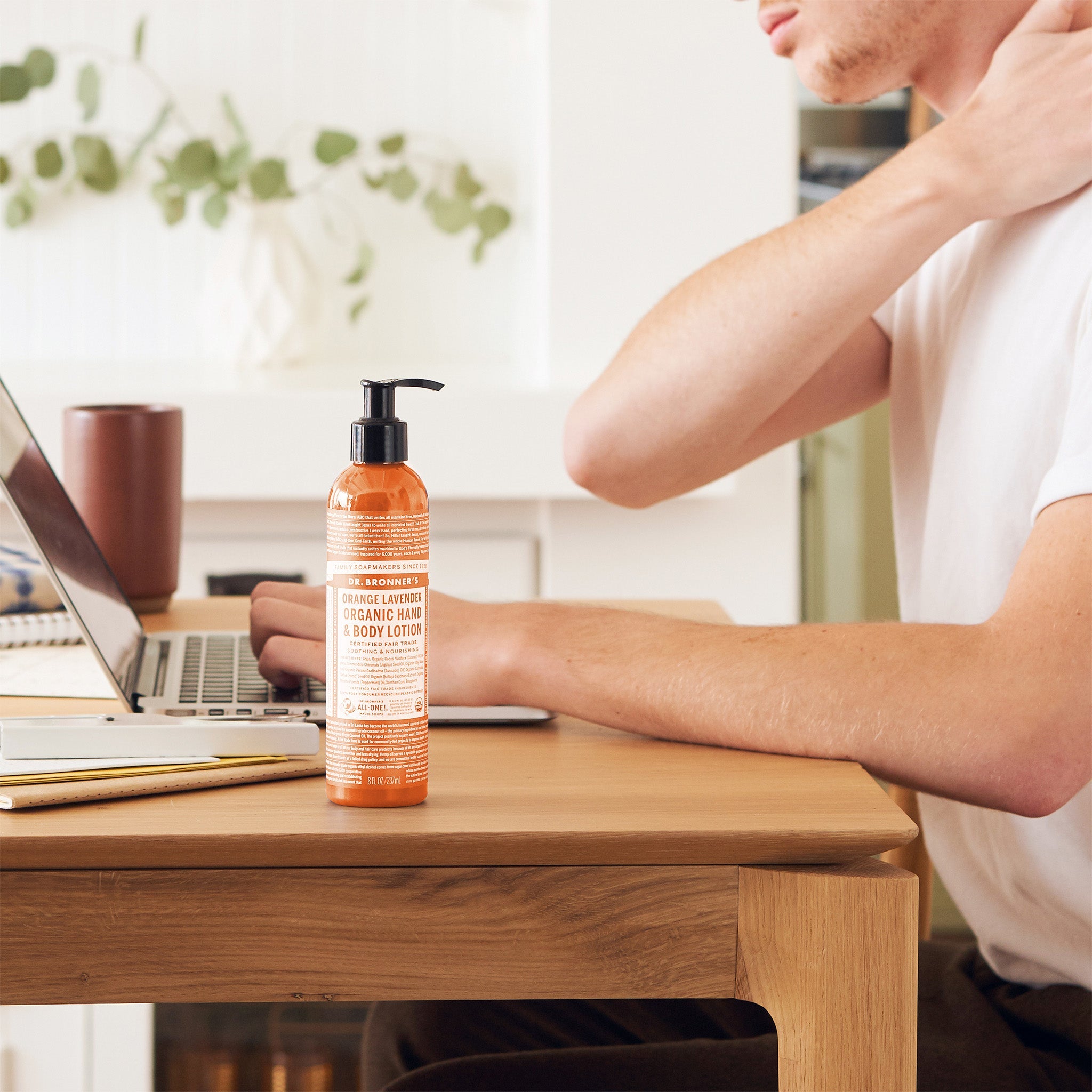 Person touching the back of their neck while seated at a wooden desk with a laptop, beside a orange Dr. Bronner's Orange Lavender Organic Hand & Body Lotion 237ml pump bottle.