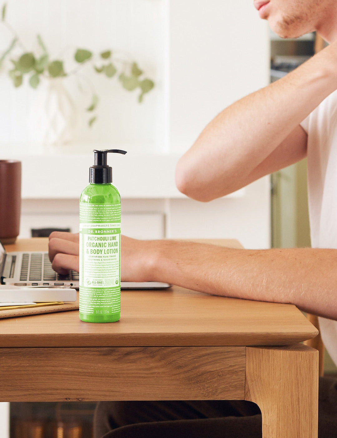 Person touching the back of their neck while seated at a wooden desk with a laptop, beside a green Dr. Bronner's Patchouli Lime Organic Hand & Body Lotion 237ml pump bottle.