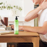 Person touching the back of their neck while seated at a wooden desk with a laptop, beside a green Dr. Bronner's Patchouli Lime Organic Hand & Body Lotion 237ml pump bottle.