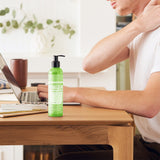 Person touching the back of their neck while seated at a wooden desk with a laptop, beside a green Dr. Bronner's Patchouli Lime Organic Hand & Body Lotion 237ml pump bottle.