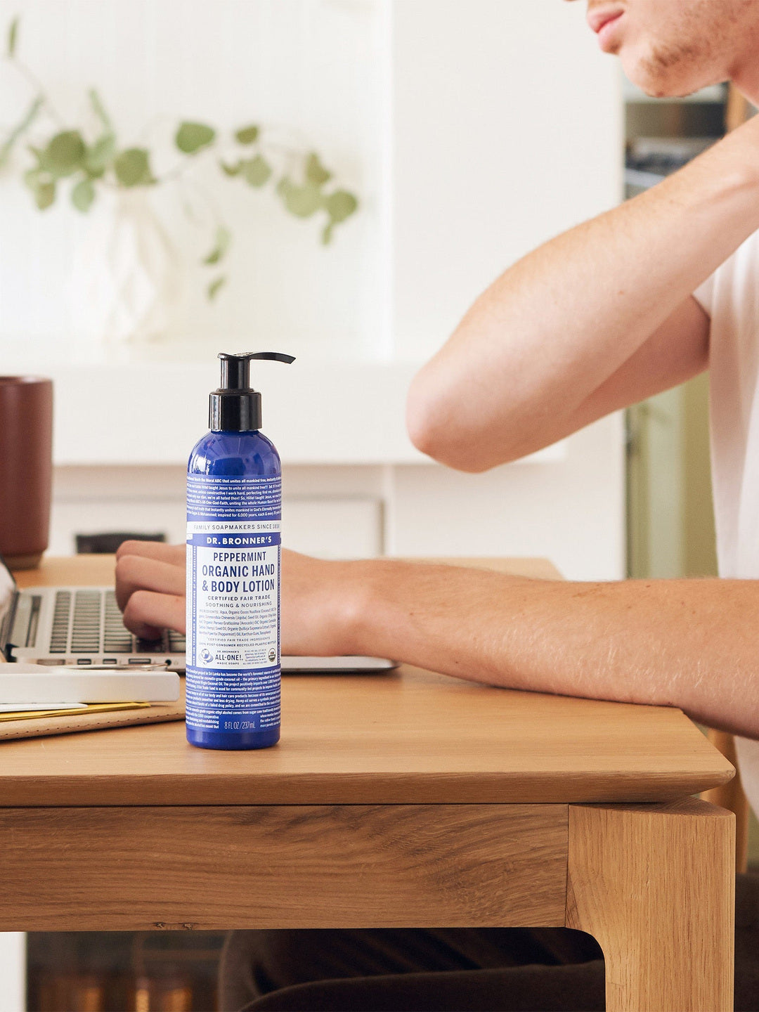 Person touching the back of their neck while seated at a wooden desk with a laptop, beside a blue Dr. Bronner's Peppermint Organic Hand & Body Lotion 237ml pump bottle.
