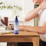 Person touching the back of their neck while seated at a wooden desk with a laptop, beside a blue Dr. Bronner's Peppermint Organic Hand & Body Lotion 237ml pump bottle.