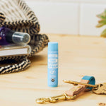 Dr. Bronner's Naked Lip Balm standing on a pale wooden counter beside keys, toiletries and a woven bag.