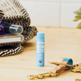 Dr. Bronner's Naked Lip Balm standing on a pale wooden counter beside keys, toiletries and a woven bag.