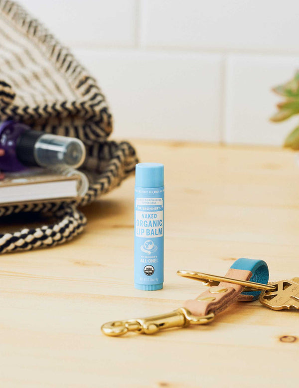 Dr. Bronner's Naked Lip Balm standing on a pale wooden counter beside keys, toiletries and a woven bag.