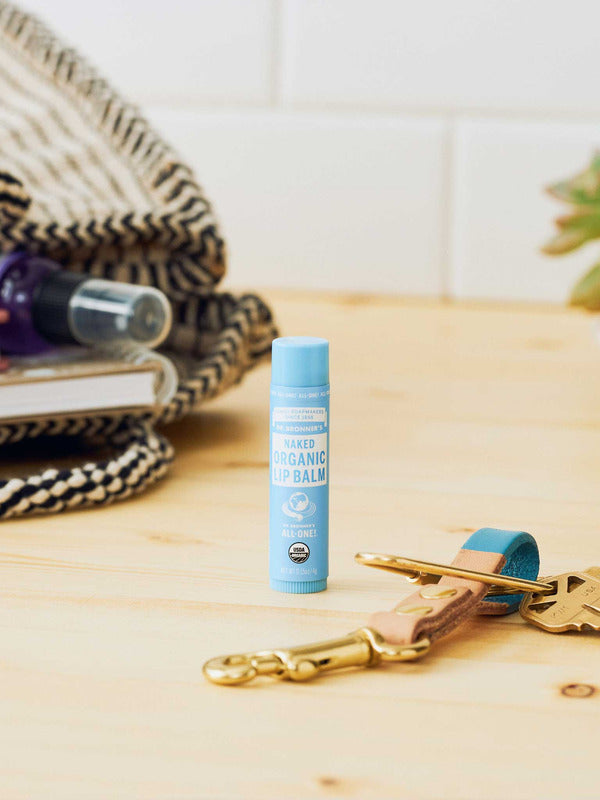 Dr. Bronner's Naked Lip Balm standing on a pale wooden counter beside keys, toiletries and a woven bag.
