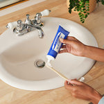 Hands squeezing blue Dr. Bronner's Peppermint Toothpaste onto a wooden toothbrush above a white bathroom basin.