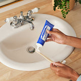 Hands squeezing blue Dr. Bronner's Peppermint Toothpaste onto a wooden toothbrush above a white bathroom basin.