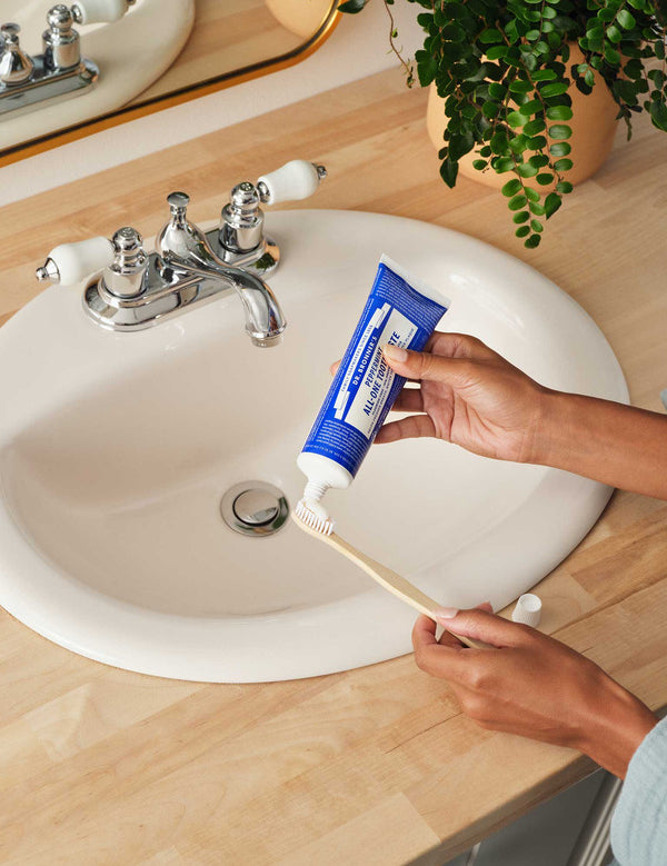 Hands squeezing blue Dr. Bronner's Peppermint Toothpaste onto a wooden toothbrush above a white bathroom basin.
