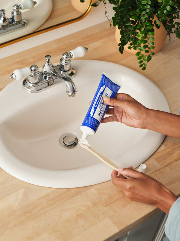 Hands squeezing blue Dr. Bronner's Peppermint Toothpaste onto a wooden toothbrush above a white bathroom basin.