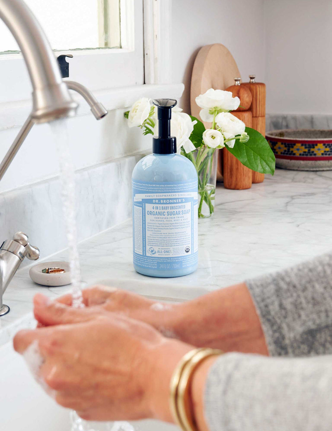 Hands lathering under running water at a white sink, with a light blue Dr. Bronner's Baby Unscented Organic Sugar Soap 710ml pump bottle on the marble bench beside white flowers.