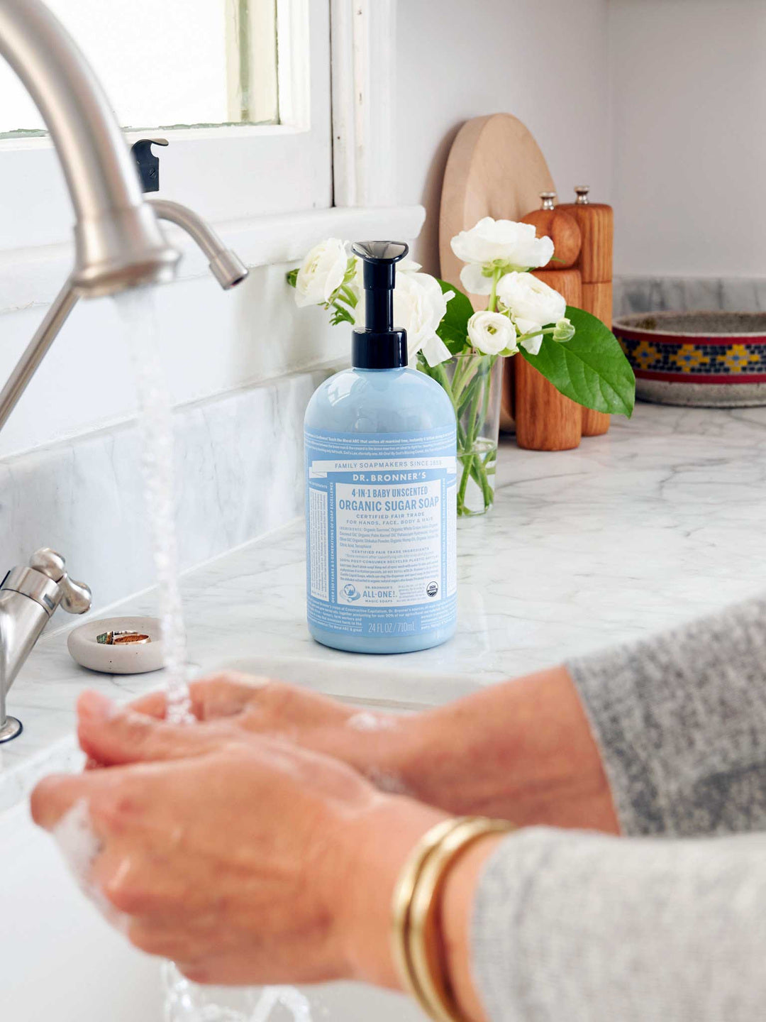 Hands lathering under running water at a white sink, with a light blue Dr. Bronner's Baby Unscented Organic Sugar Soap 710ml pump bottle on the marble bench beside white flowers.
