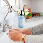 Hands lathering under running water at a white sink, with a light blue Dr. Bronner's Baby Unscented Organic Sugar Soap 710ml pump bottle on the marble bench beside white flowers.