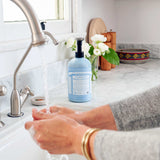 Hands lathering under running water at a white sink, with a light blue Dr. Bronner's Baby Unscented Organic Sugar Soap 710ml pump bottle on the marble bench beside white flowers.