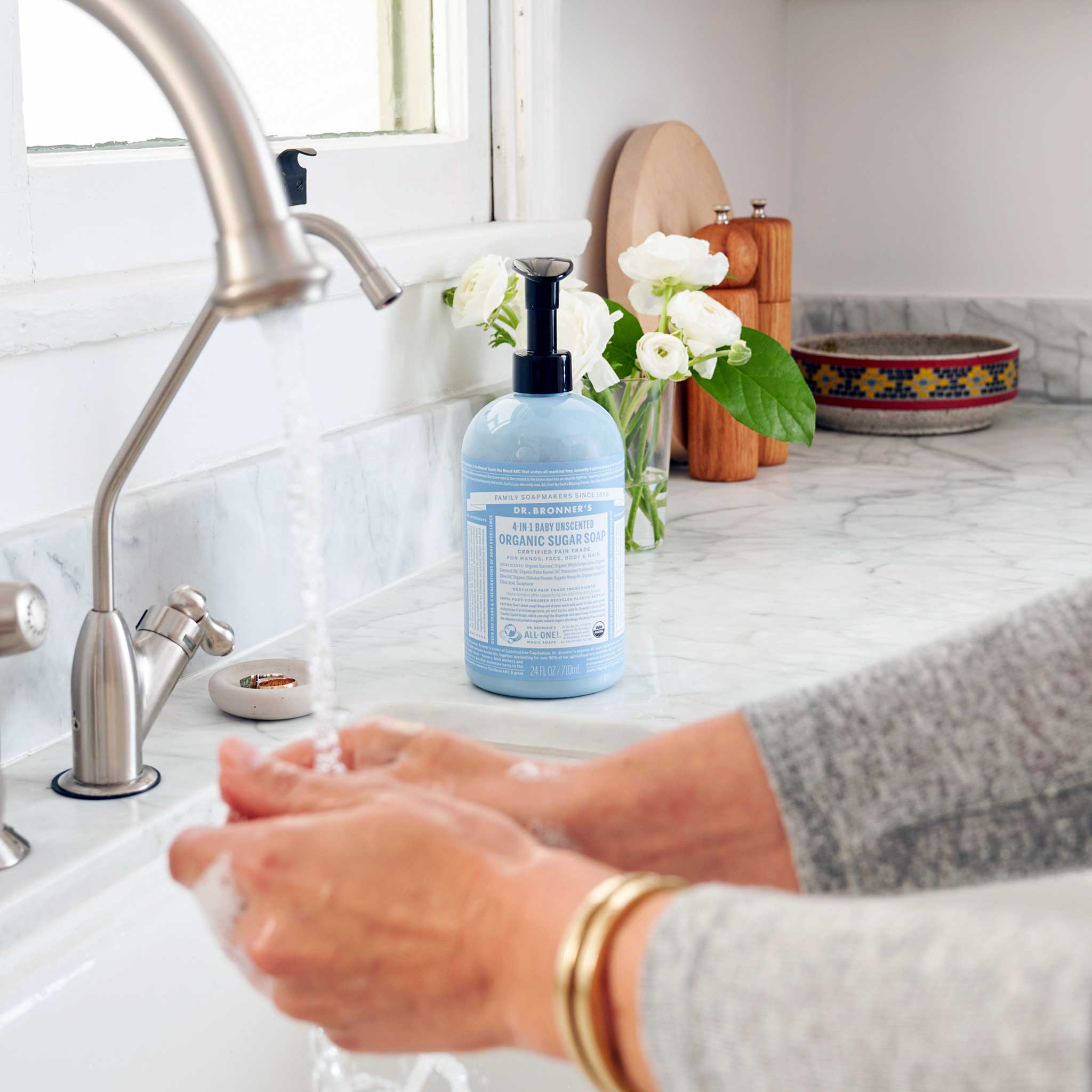 Hands lathering under running water at a white sink, with a light blue Dr. Bronner's Baby Unscented Organic Sugar Soap 710ml pump bottle on the marble bench beside white flowers.