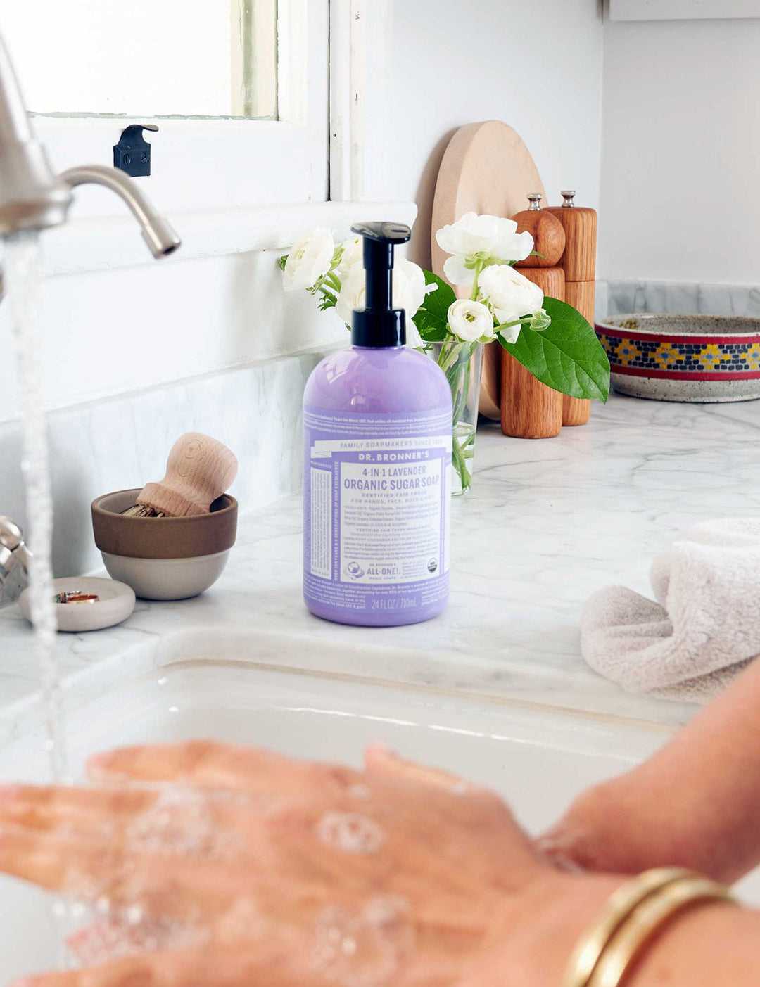 Hands washing with lather at a kitchen sink beside a purple Dr. Bronner's Lavender Organic Sugar Soap 710ml pump bottle, a folded towel and white flowers.