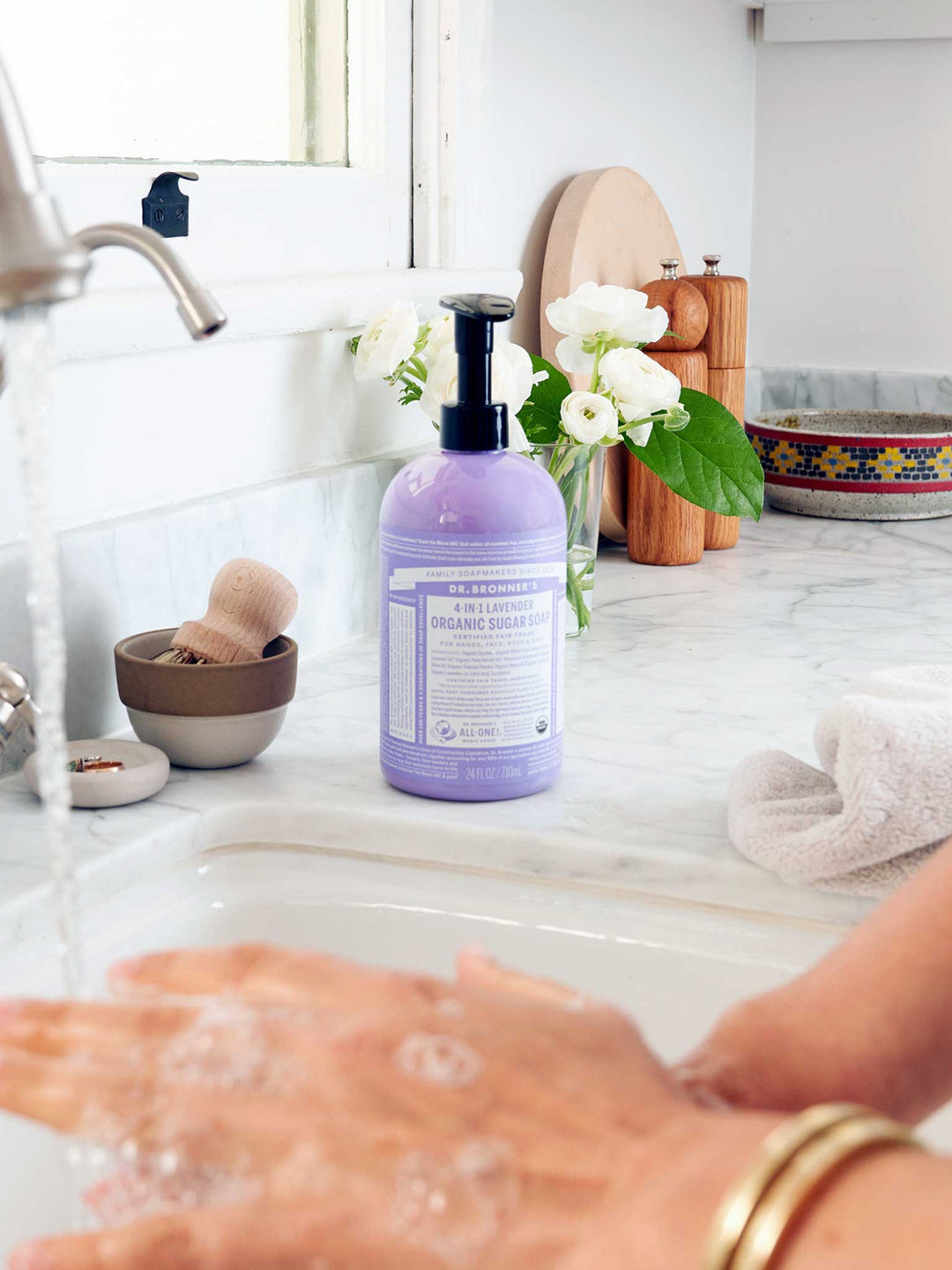 Hands washing with lather at a kitchen sink beside a purple Dr. Bronner's Lavender Organic Sugar Soap 710ml pump bottle, a folded towel and white flowers.