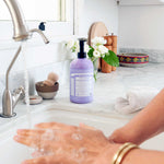 Hands washing with lather at a kitchen sink beside a purple Dr. Bronner's Lavender Organic Sugar Soap 710ml pump bottle, a folded towel and white flowers.