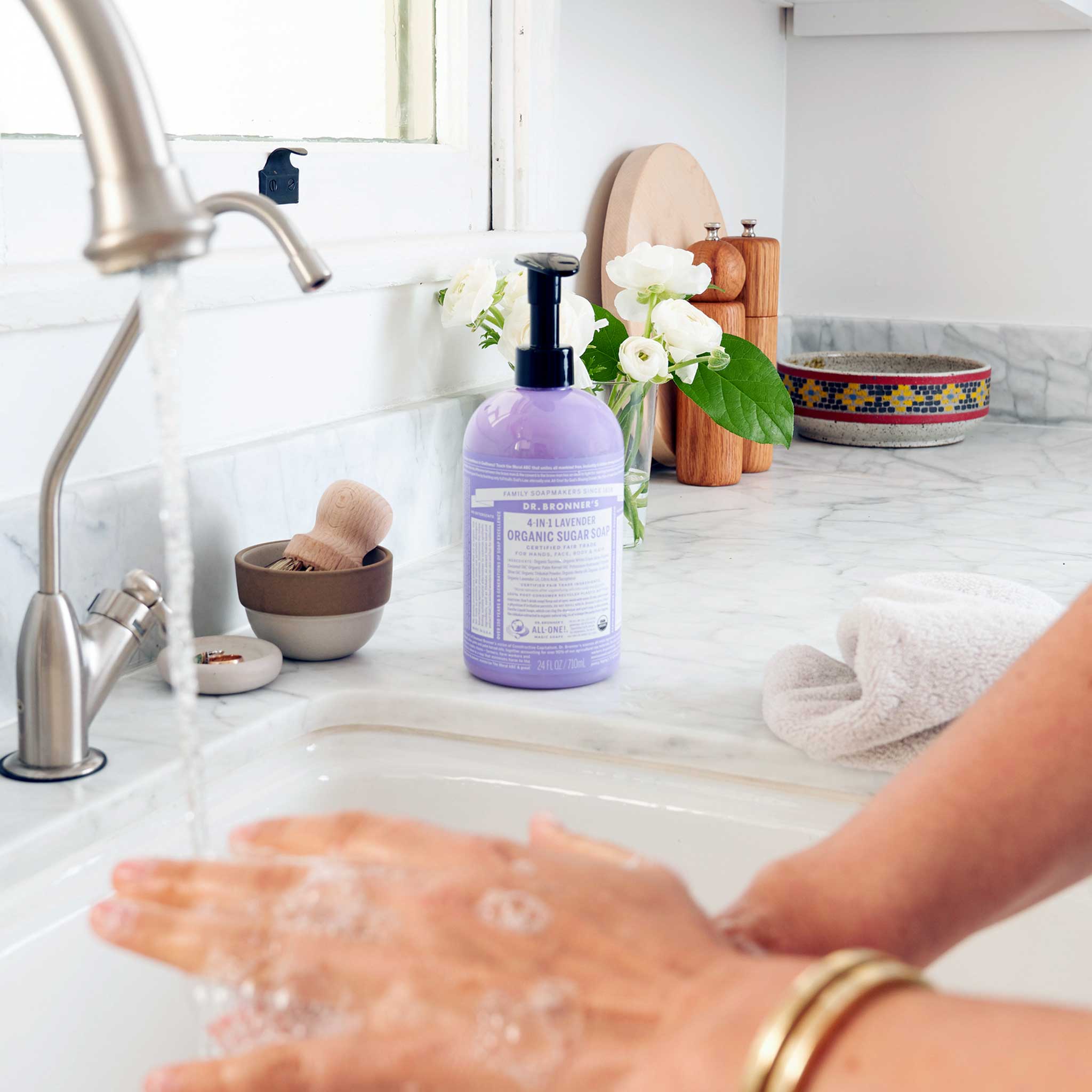 Hands washing with lather at a kitchen sink beside a purple Dr. Bronner's Lavender Organic Sugar Soap 710ml pump bottle, a folded towel and white flowers.