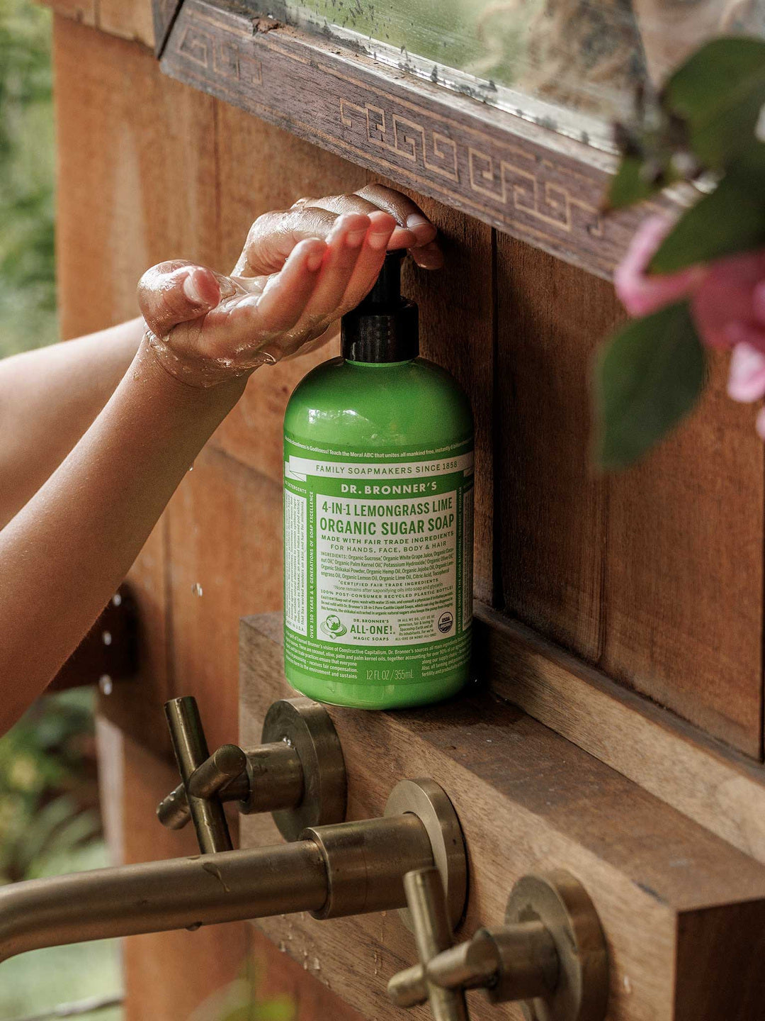 Hands pressing the black pump of a green Dr. Bronner's Lemongrass Lime Organic Sugar Soap 355ml bottle at a wooden washstand with brass taps.