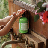 Hands pressing the black pump of a green Dr. Bronner's Lemongrass Lime Organic Sugar Soap 355ml bottle at a wooden washstand with brass taps.