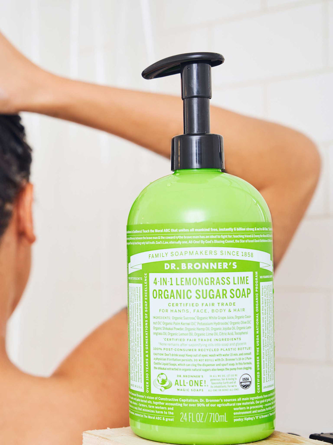 Rear view of a person lathering their wet hair in the shower, beside a green Dr. Bronner's Lemongrass Lime Organic Sugar Soap 710ml pump bottle in the foreground.