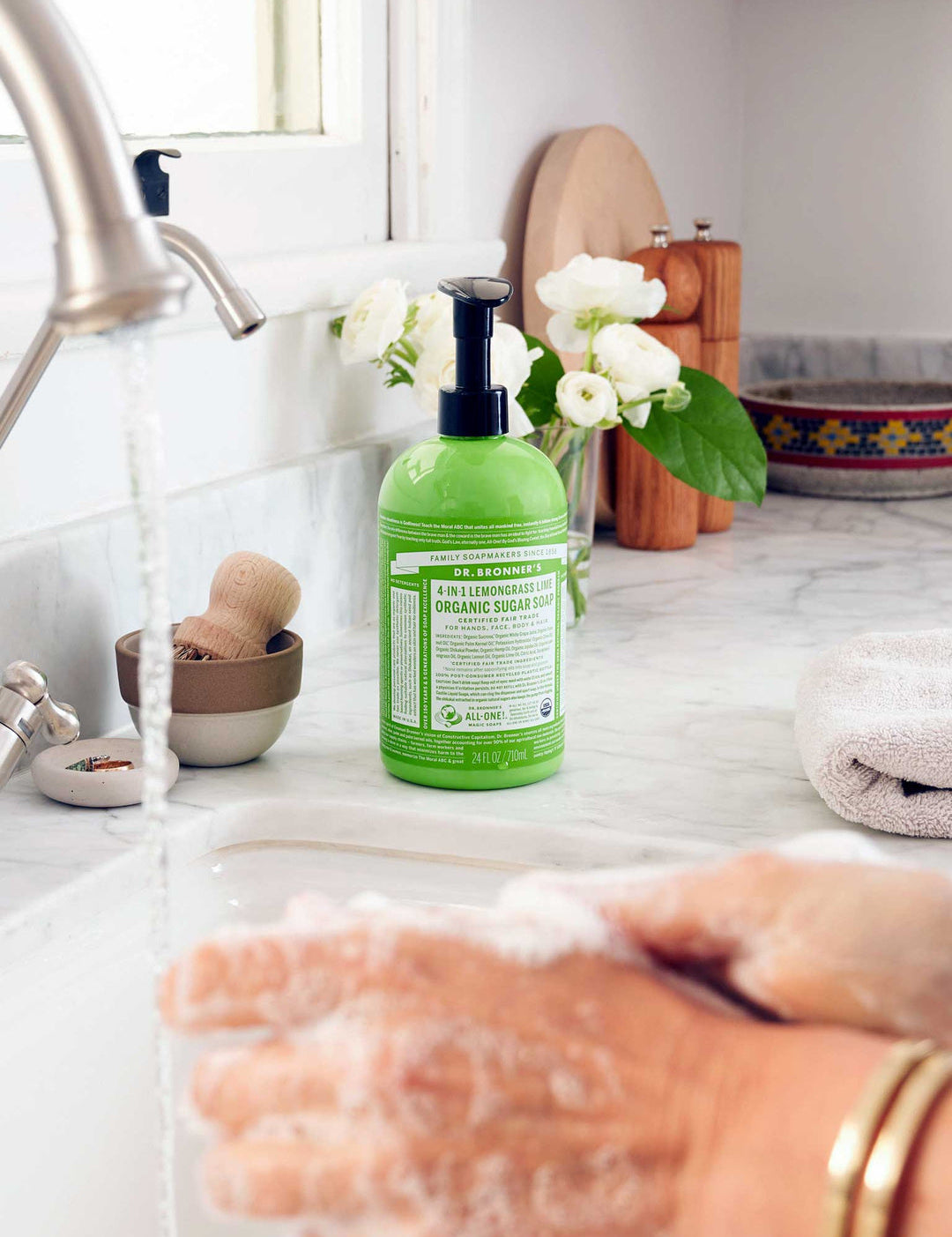 Hands rubbing together with soap lather over a white sink, with a green Dr. Bronner's Lemongrass Lime Organic Sugar Soap 710ml pump bottle standing on the marble bench.