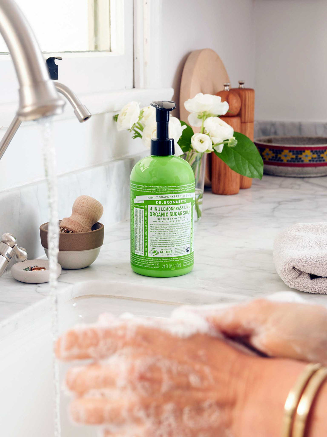 Hands rubbing together with soap lather over a white sink, with a green Dr. Bronner's Lemongrass Lime Organic Sugar Soap 710ml pump bottle standing on the marble bench.