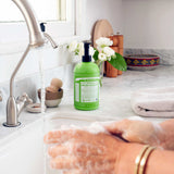 Hands rubbing together with soap lather over a white sink, with a green Dr. Bronner's Lemongrass Lime Organic Sugar Soap 710ml pump bottle standing on the marble bench.