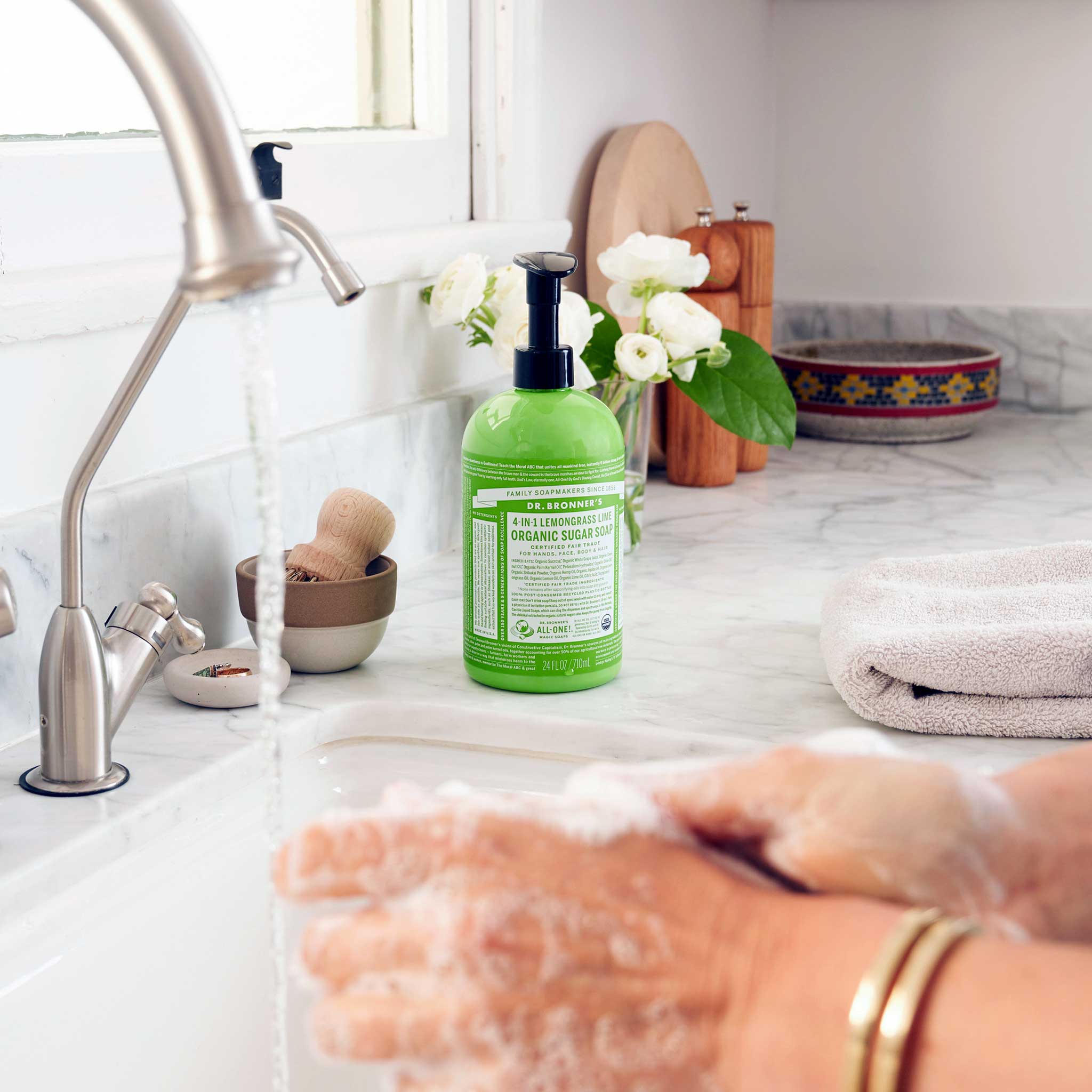 Hands rubbing together with soap lather over a white sink, with a green Dr. Bronner's Lemongrass Lime Organic Sugar Soap 710ml pump bottle standing on the marble bench.