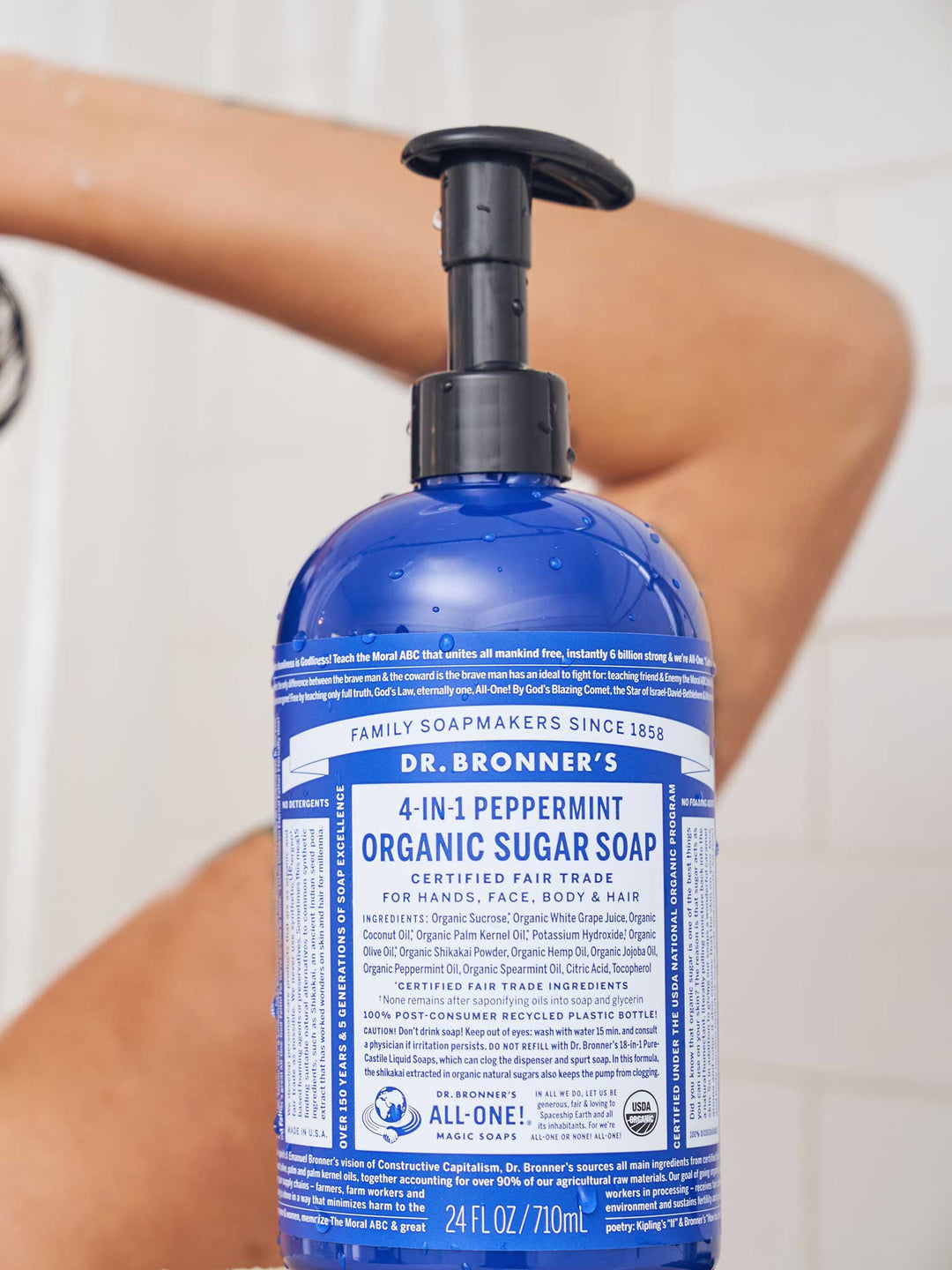 Rear view of a person washing their hair in the shower, with a blue Dr. Bronner's Peppermint Organic Sugar Soap 710ml pump bottle clearly visible in the foreground.