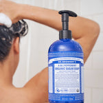 Rear view of a person washing their hair in the shower, with a blue Dr. Bronner's Peppermint Organic Sugar Soap 710ml pump bottle clearly visible in the foreground.