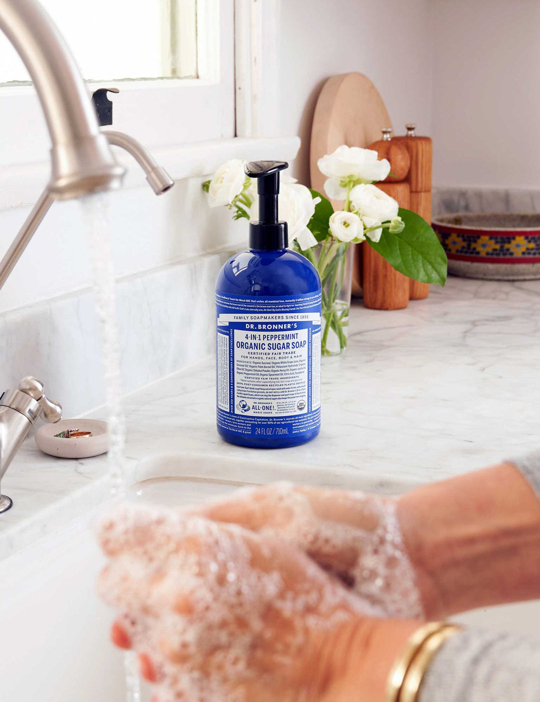 Hands washing with white soap lather over a sink, beside a blue Dr. Bronner's Peppermint Organic Sugar Soap 710ml pump bottle on a marble bench.