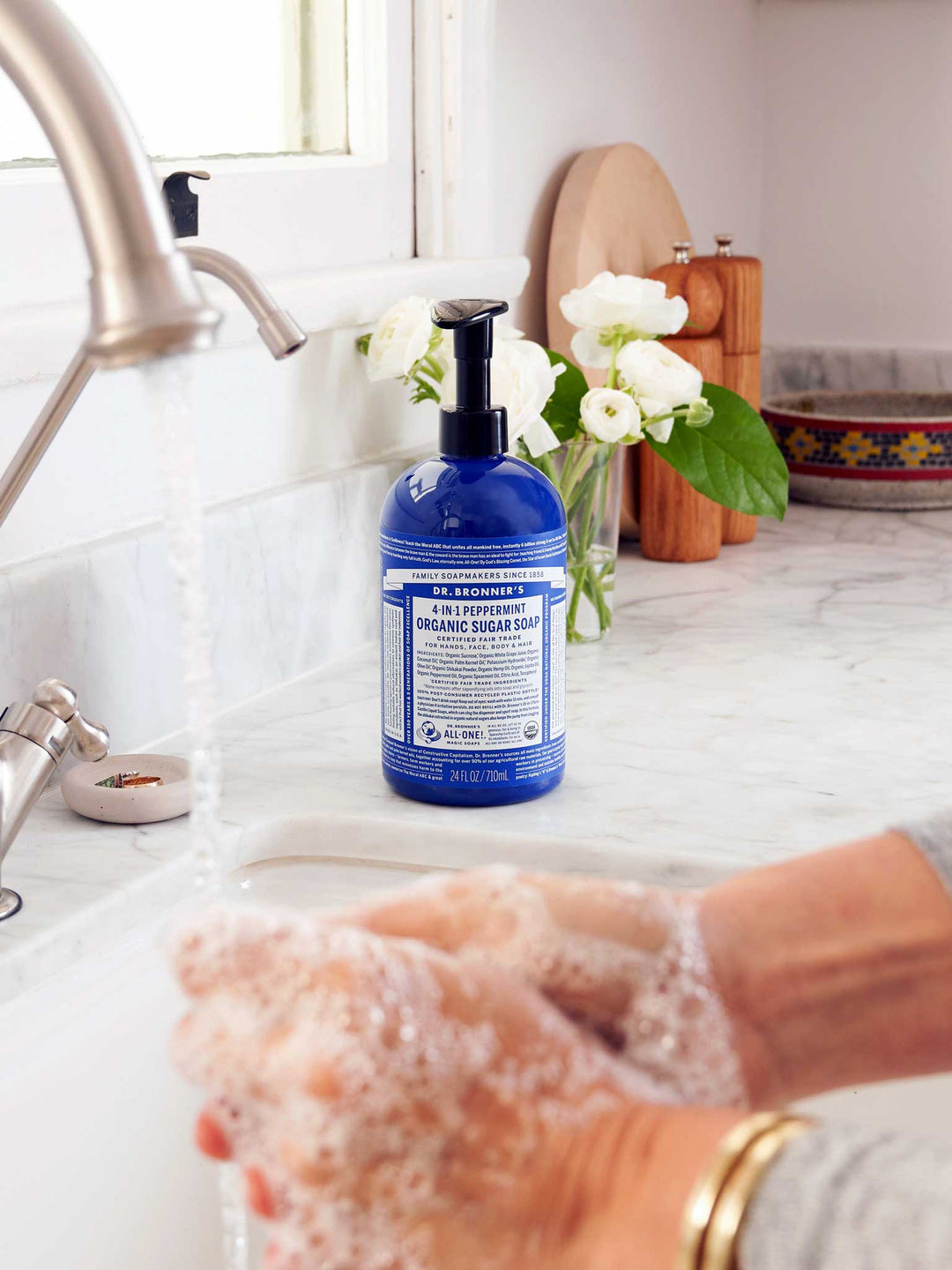 Hands washing with white soap lather over a sink, beside a blue Dr. Bronner's Peppermint Organic Sugar Soap 710ml pump bottle on a marble bench.